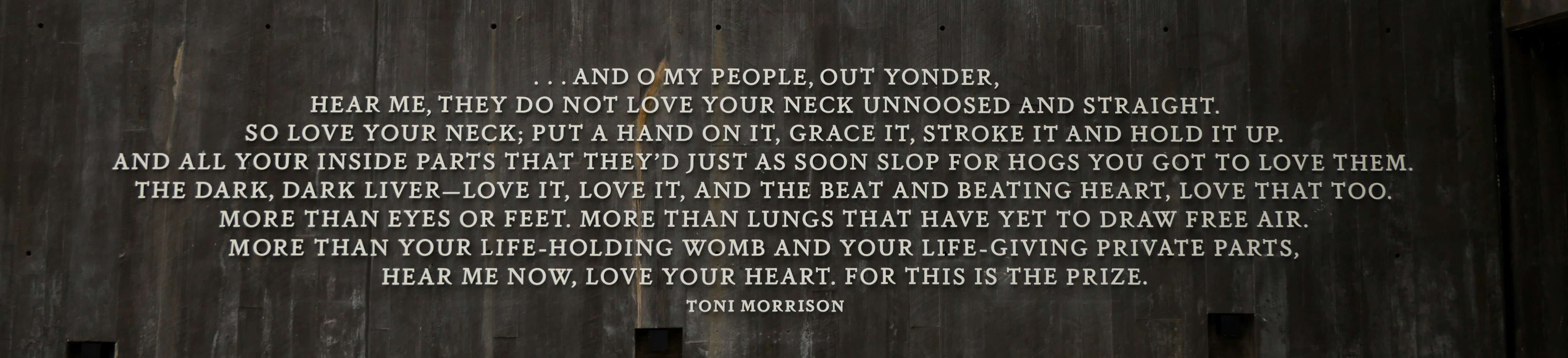 A quote engraved in white text on nearly black marble:  '… AND O MY PEOPLE, OUT YONDER, HEAR ME, THEY DO NOT LOVE YOUR NECK UNNOOSED AND STRAIGHT. SO LOVE YOUR NECK; PUT A HAND ON IT, GRACE IT, STROKE IT AND HOLD IT UP. AND ALL YOUR INSIDE PART'S THAT THEY'D JUST AS SOON SLOP FOR HOGS YOU GOT TO LOVE THEM. THE DARK, DARK LIVER - LOVE IT, LOVE IT, AND THE BEAT AND BEATING HEART, LOVE THAT TOO. MORE THAN EYES OR FEET. MORE THAN LUNGS THAT HAVE YET TO DRAW FREE AIR. MORE THAN YOUR LIFE-HOLDING WOMB AND YOUR LIFE-GIVING PRIVATE PARTS, HEAR ME NOW, LOVE YOUR HEART. FOR THIS IS THE PRIZE.' - Toni Morrison